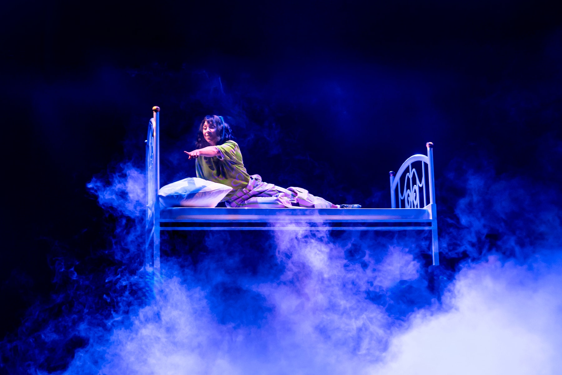 Luna ligt op een zwevend bed, omringd door blauwe verlichting en mist, terwijl ze met een uitgestrekte arm naar voren wijst tijdens de illusieshow Magicaluna in het Efteling Theater.