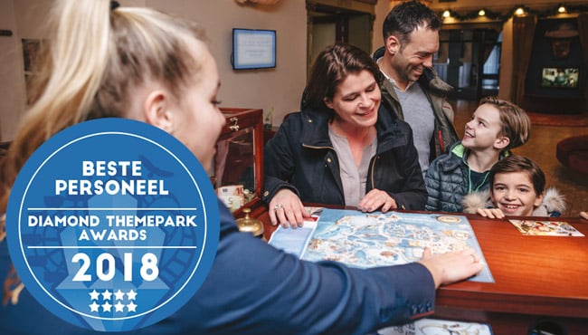 Foto van een medewerkster van de Efteling, achter de balie van het Efteling Hotel. Ze laat een parkplattegrond zien van de Efteling aan een gezin. Verder staat er een button voor het beste personeel op de foto.