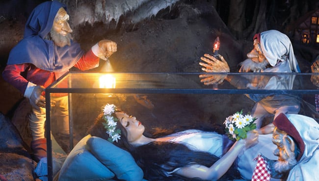 Sneeuwwitje in haar glazen kist met drie van de zeven dwergen om haar heen.