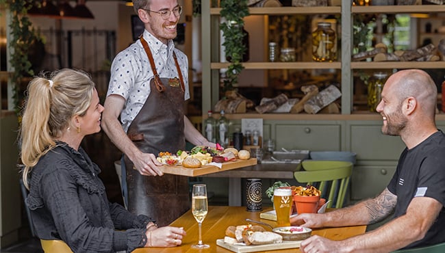 Medewerker serveert een plank met eten aan twee gasten in Restaurant de Proeftuyn.