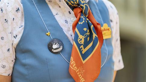 Een Efteling-medewerker draagt een blauwe button met een handgebaar als symbool voor gebarentaalvaardigheid op een blauw uniform met oranje halsdoek.
